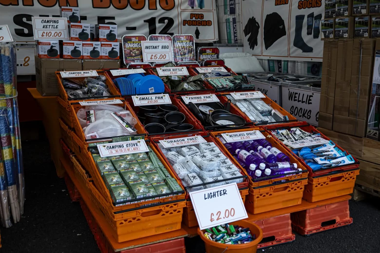 Crates of camping cookware under handwritten price cards: kettles, mugs, pans, mess tins, stoves, fuel blocks and lighters.